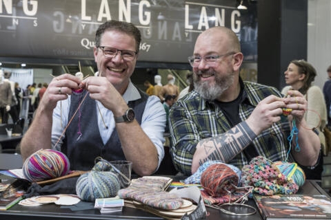 two men smiling and knitting at a table